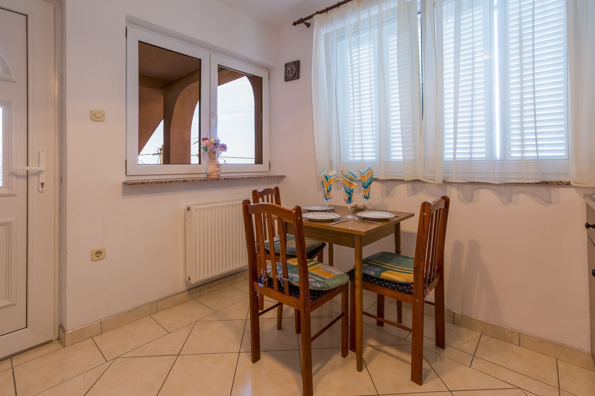 A dining area with a wooden table and four chairs, a window with blinds, a radiator, and a clock on the wall.