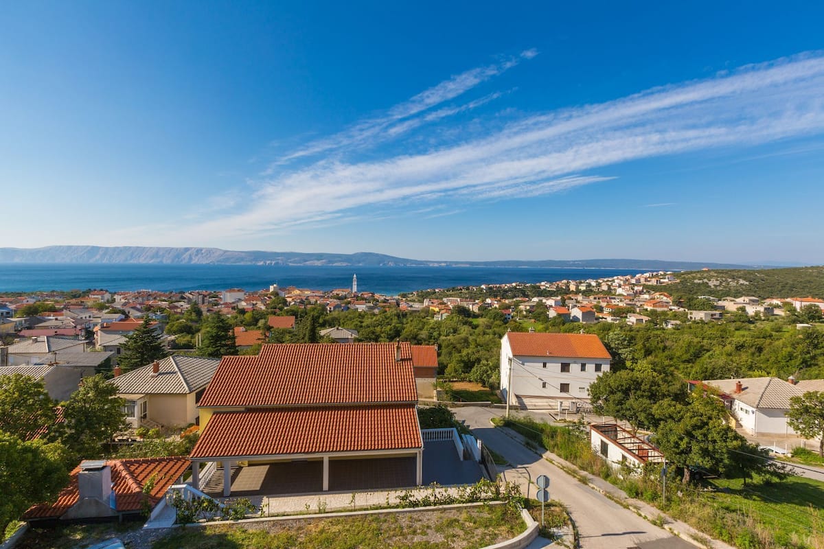 Eine Küstenstadt mit Häusern mit Terrakotta-Dächern, blauem Himmel und einer weiten Aussicht auf Meer und Berge.