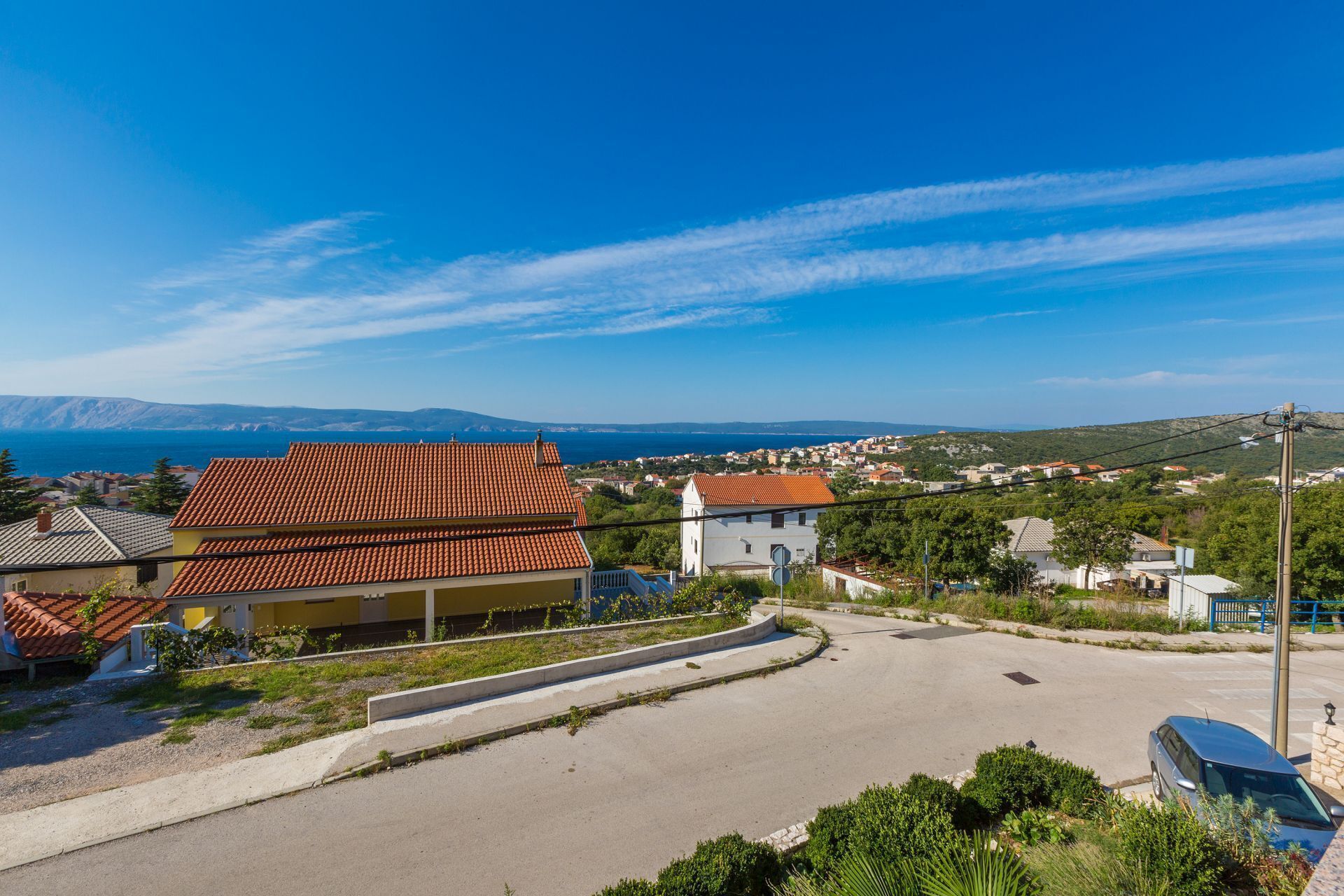 Ein Küstenwohngebiet mit Häusern mit Terrakotta-Dächern, blauem Himmel und Blick auf das Meer und die entfernten Hügel.