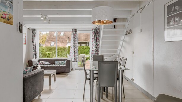 A modern living room with a sofa, dining table, and chairs. There is a staircase leading to an upper level.