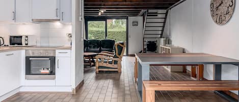 A kitchen with white cabinets, a microwave, and an oven. There is a wooden bench and a table with a clock on the wall.