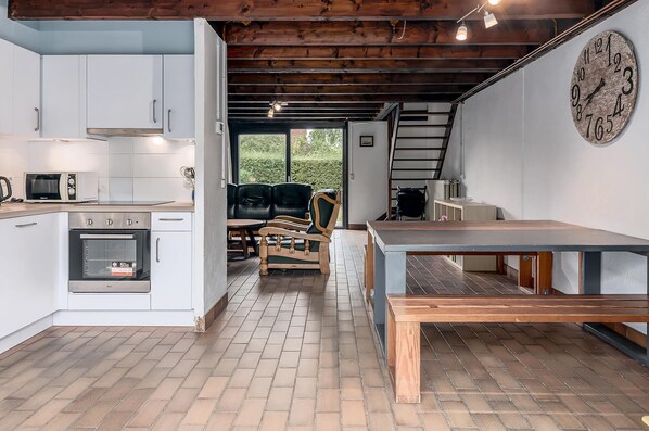 A kitchen with white cabinets, a microwave, and an oven. There is a wooden bench and a table with a clock on the wall.