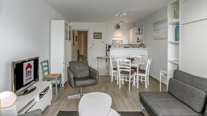 Ett modernt vardagsrum med en grå soffa, ett vitt matbord med stolar, en grå fåtölj och en tv-skärm som visar en logotyp för ett fastighetsbolag.