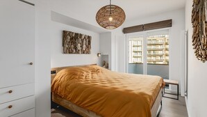 Une chambre à coucher comprenant un lit, une commode blanche, une tête de lit en bois, une œuvre d’art accrochée au mur, un luminaire suspendu et une fenêtre donnant sur des immeubles.