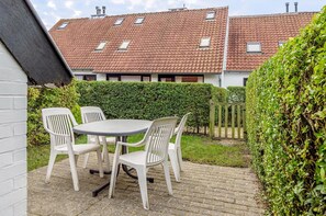 Een terras met een ronde tafel en vier witte stoelen, omgeven door een haag en een houten omheining.