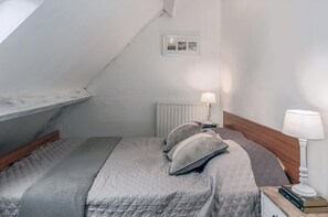 Een slaapkamer met een schuine kap, een bed met grijze beddengoed, twee nachtlampjes, een radiator en een klein fotolijstje aan de muur.