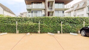 Un estacionamiento con suelo de arena, un seto y un edificio de varios pisos con balcones al fondo.