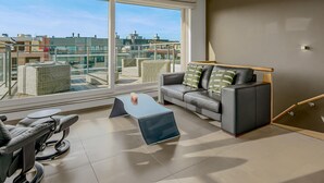 A modern living room with a large window, a grey sofa, a massage chair, and a small coffee table.