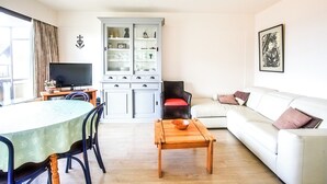 Ein Wohnzimmer mit einem weißen Sofa, einem Holztisch, einem Esstisch mit Stühlen, einem Fernseher und einer Vitrine mit Glastüren.
