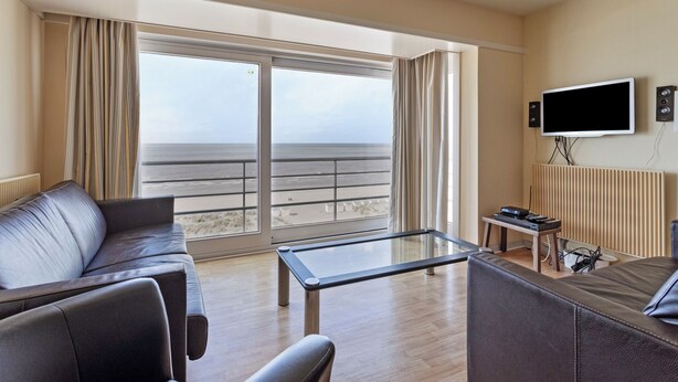 Una sala de estar moderna con un amplio ventanal con vista a la playa, un televisor de pantalla plana montado en la pared y una pequeña mesa con dispositivos electrónicos.