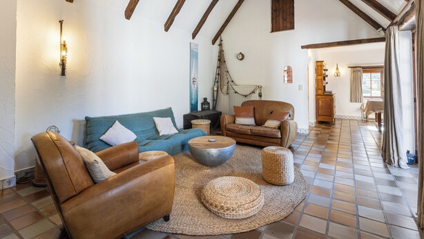 Amplia sala de estar con techo alto, vigas de madera a la vista, un ventanal grande y una zona de estar acogedora con sofá, sillón y reposapiés.