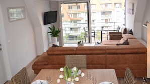 Un salon moderne avec un canapé marron, une table à manger dressée avec de l’assiette et des verres blancs, et une grande fenêtre donnant sur un paysage urbain.
