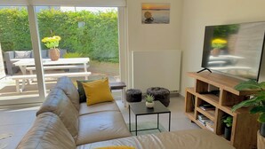 A living room with a beige sofa, a TV on a wooden stand, and a view of a garden through a sliding glass door. - 2 bedroom accommodation in Oostduinkerke (Oostduinkerke)