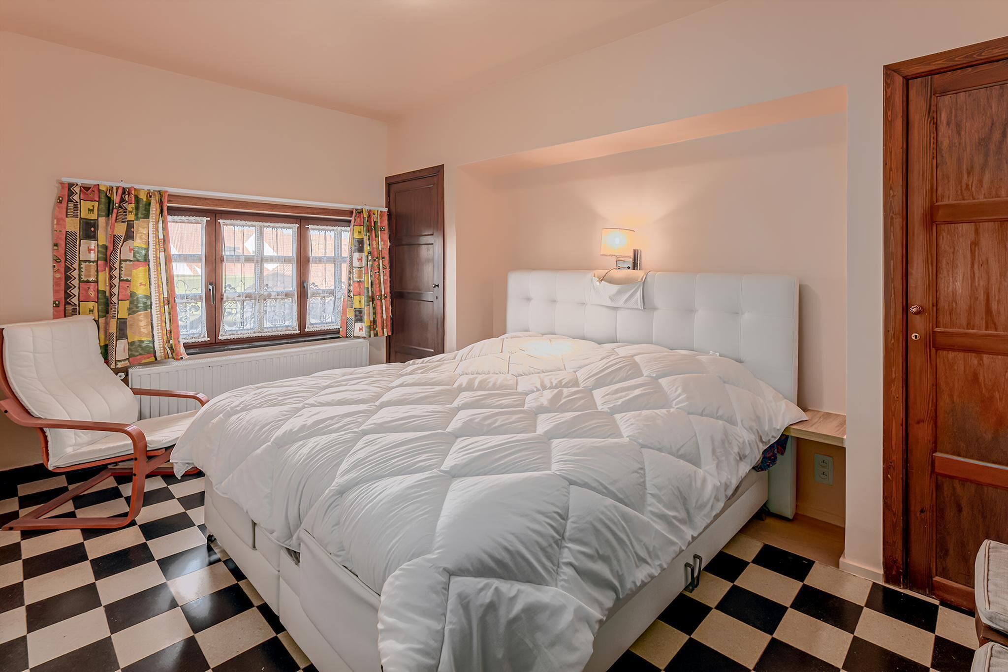 Una camera da letto con pavimento a scacchi, un letto bianco con testiera capitonata, una sedia bianca e una finestra con tende colorate.