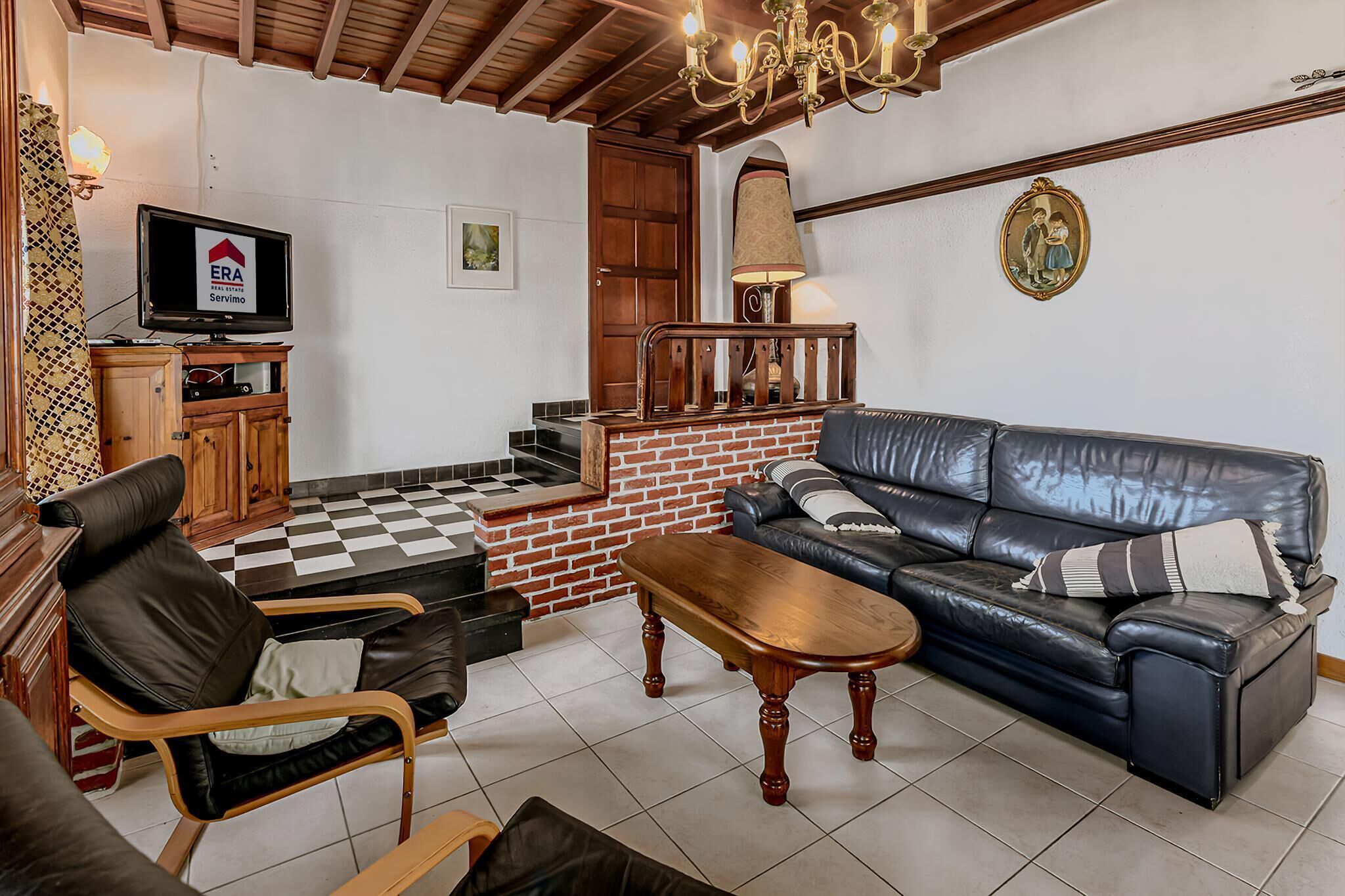 Un salon avec un sol à carreaux, un canapé, une table basse en bois, un fauteuil en cuir, un téléviseur posé sur un meuble en bois et un escalier avec un mur en briques rouges.