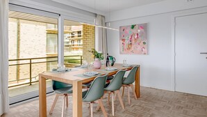 A dining area with a wooden table set for four, green chairs, and a view of a balcony with a brick building. - 2 bedroom accommodation in Oostduinkerke (Oostduinkerke)