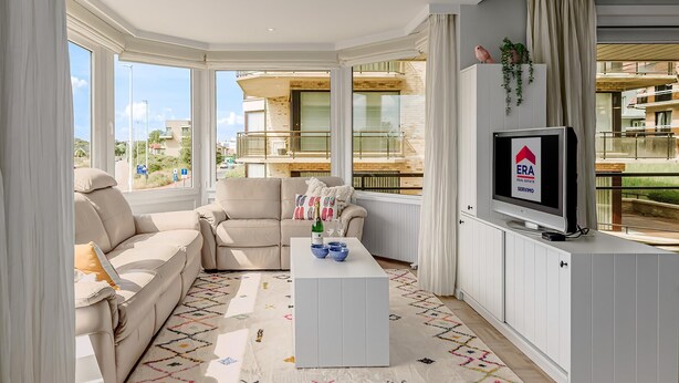 A modern living room with a sofa, armchair, and a coffee table. There is a TV on a cabinet, and large windows offer a view of the outside. - 2 bedroom accommodation in Oostduinkerke (Oostduinkerke)