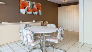 Un coin repas avec une table ronde et des chaises colorées, une cuisine avec des meubles de couleur beige et un mur orné de deux tableaux représentant des fleurs.