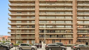 Un immeuble d’appartements à plusieurs étages, doté d’un ascenseur central, de nombreux balcons et d’un espace de stationnement où sont garées plusieurs voitures.