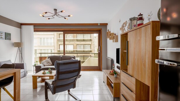 Ein modernes Wohnzimmer mit einer Couch, einem Couchtisch und einem Fernseher auf einer Holzkommode. Ein Balkon bietet Blick auf Gebäude.