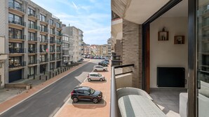 Ein modernes Mehrfamilienhaus mit mehreren Balkonen, eine Straßenansicht mit parkenden Autos und Blick auf Nachbargebäude.