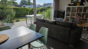 A modern living room with a dark sofa, a TV, and a bookshelf. There is a dining area with a table and chairs. The room has large windows with a view of a garden and buildings. - 2 bedroom accommodation in Oostduinkerke (Oostduinkerke)