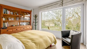 Ein Schlafzimmer mit einem großen Bett, einer Holzregal, einem schwarzen Sessel und einem großen Fenster mit Blick auf Bäume und Häuser.