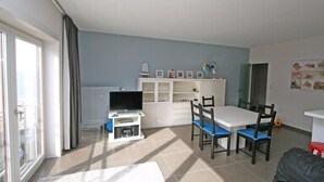 A living room with a TV, white cabinets, a dining table with chairs, and a view through a glass door. - WESTDIEP DOM.0502+G1074 (Nieuwpoort)