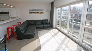 A modern living room with a black sectional sofa, red bar stools, a large window with a view of buildings, and a kitchen area with white cabinets. - WESTDIEP DOM.0502+G1074 (Nieuwpoort)