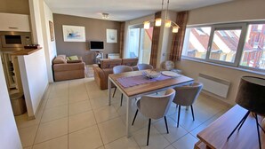 A modern living room with a dining area, a flat-screen TV, and a large window with a view of buildings. - 2 bedroom accommodation in Nieuwpoort (Nieuwpoort)