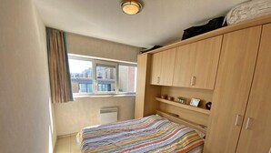 A bedroom with a bed, a window, a radiator, and wooden cabinets. - 2 bedroom accommodation in Nieuwpoort (Nieuwpoort)