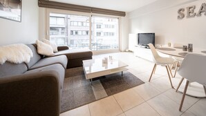 A modern living room with a sofa, coffee table, and dining area with chairs. A television is mounted on the wall, and there is a large window with a view of buildings outside.