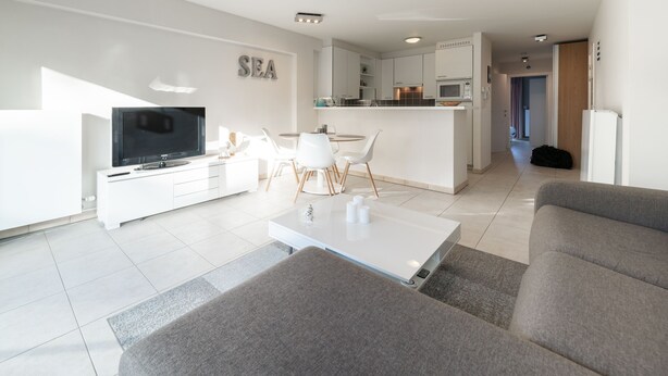 A modern living room with a flat-screen TV, a dining area with a white table and chairs, and a kitchen in the background.