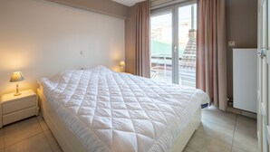 A neatly made bed in a hotel room with a nightstand, lamp, and a view of the outside through a sliding door.