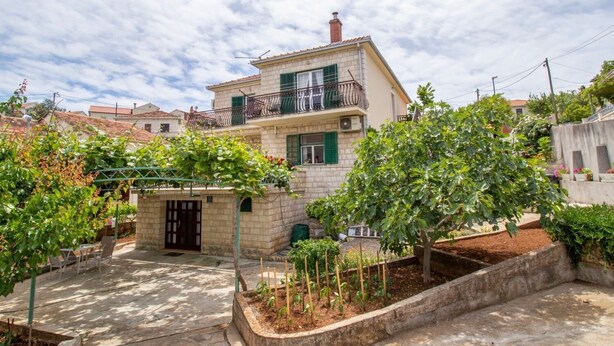 A two-story house with a balcony, a small garden area with trees and plants, and a paved driveway. - Apartment Zvone (Supetar)