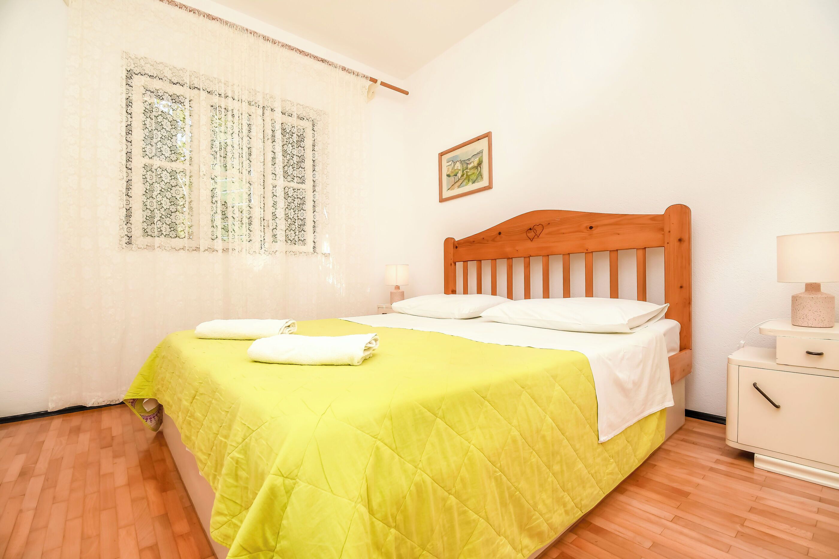 A neatly made bed with a yellow bedspread and white pillows, a wooden headboard, a bedside table with a lamp, and a window with patterned curtains.