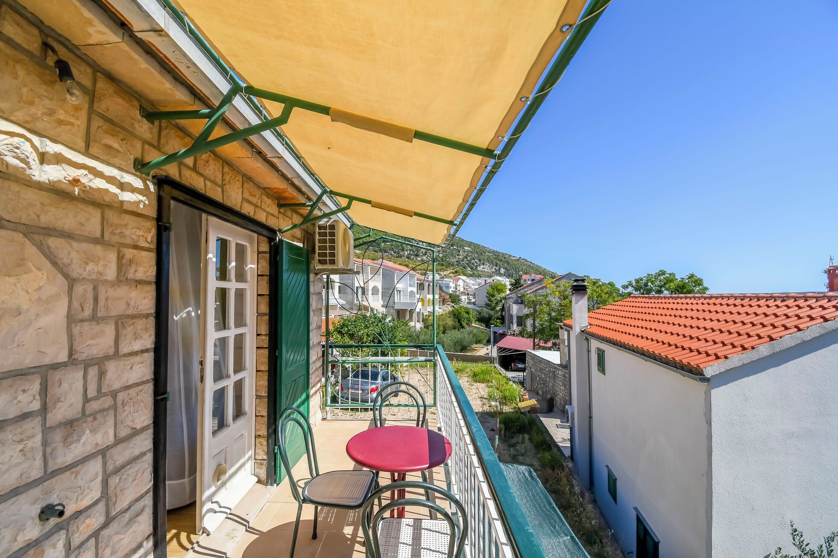 En balkon med et rødt bord og stole, en gul markise og udsigt til en skråning med bygninger.