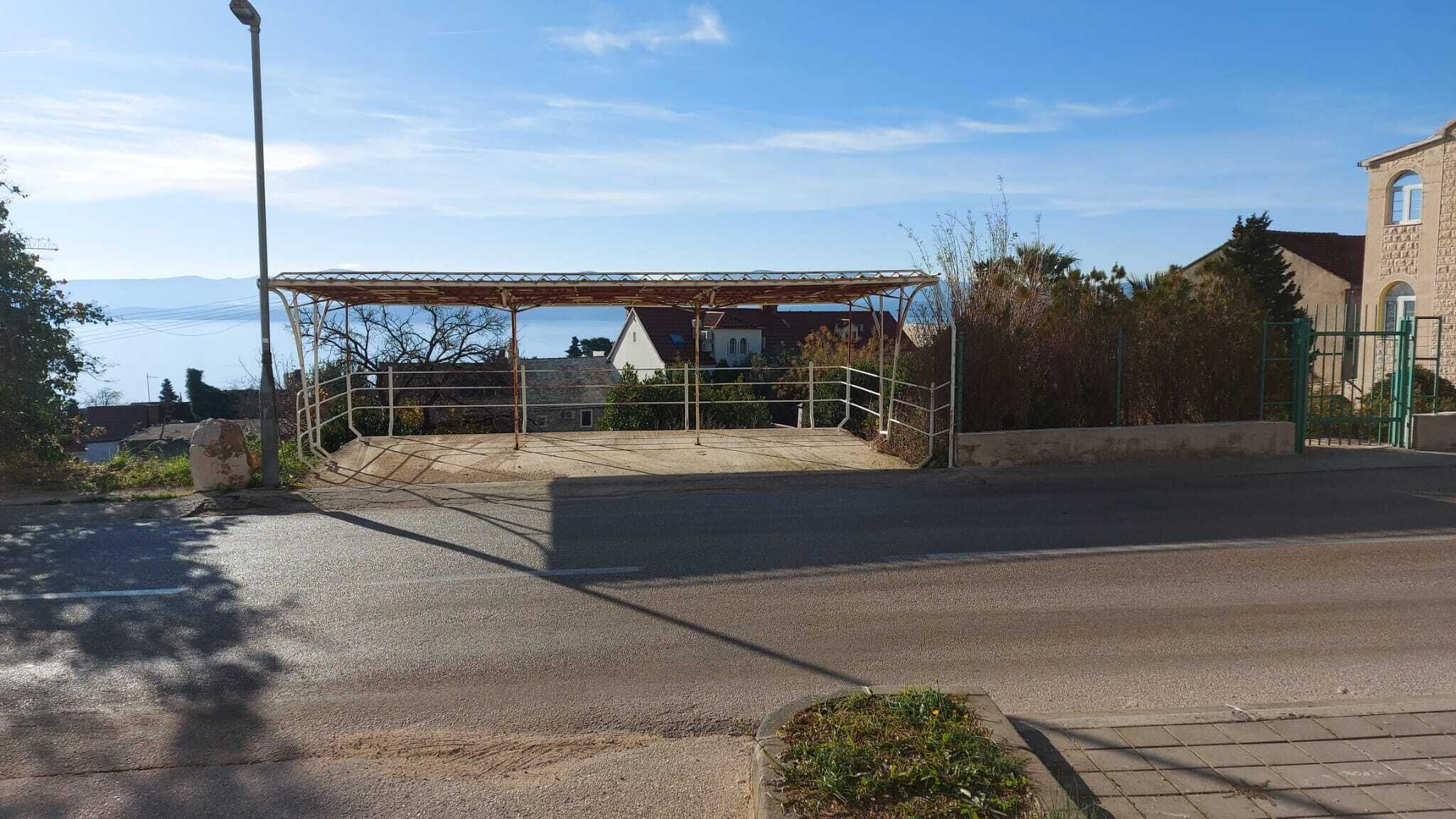 Una strada asfaltata che conduce a un'area di parcheggio coperta, con un cancello in metallo, un lampione e un edificio con facciata in pietra sullo sfondo.
