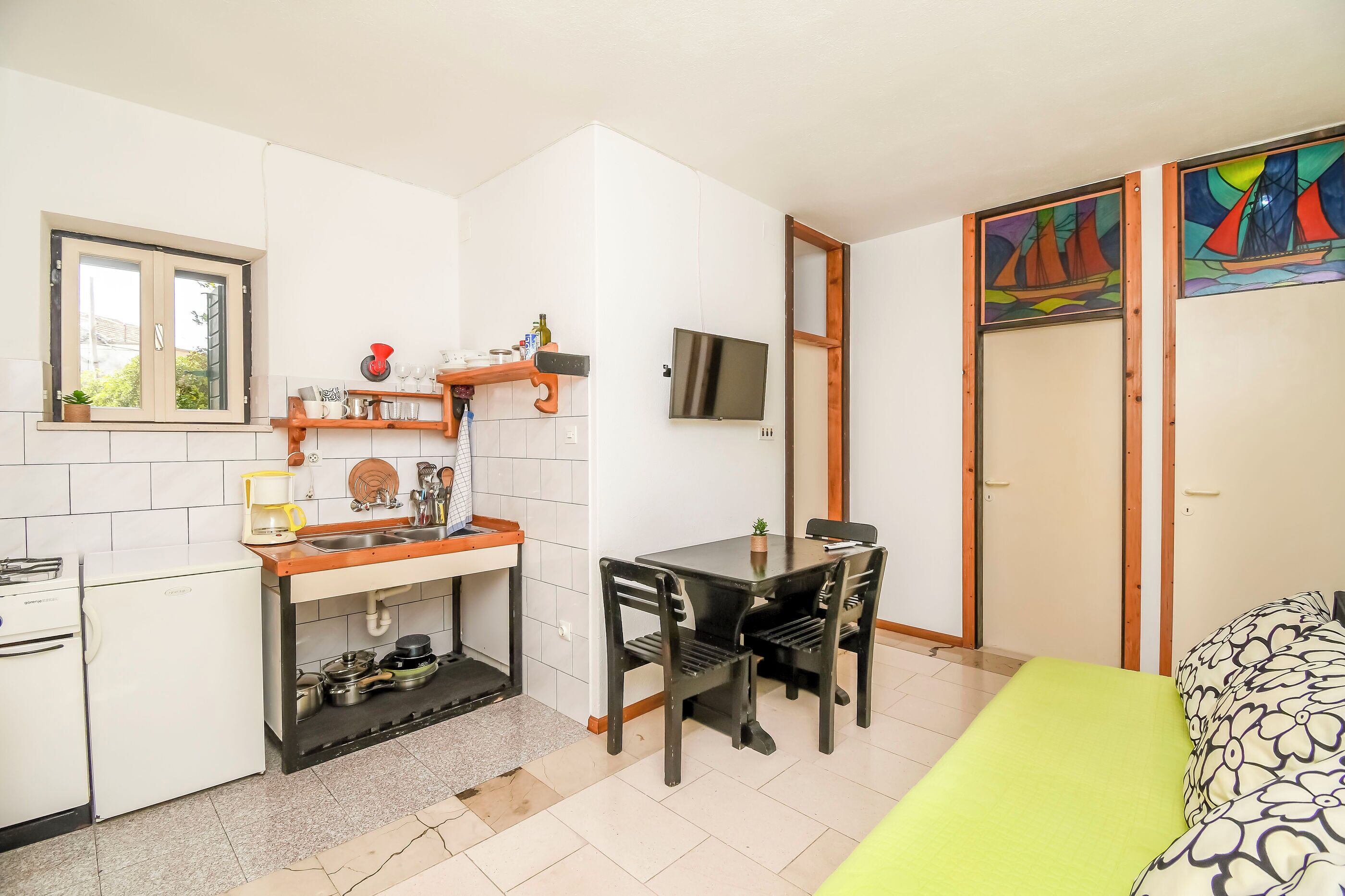 Een compacte keuken met een spoelbak, fornuis en een eethoek met stoelen. Een televisie is aan de muur bevestigd, en er zijn twee deuren die naar andere kamers leiden.