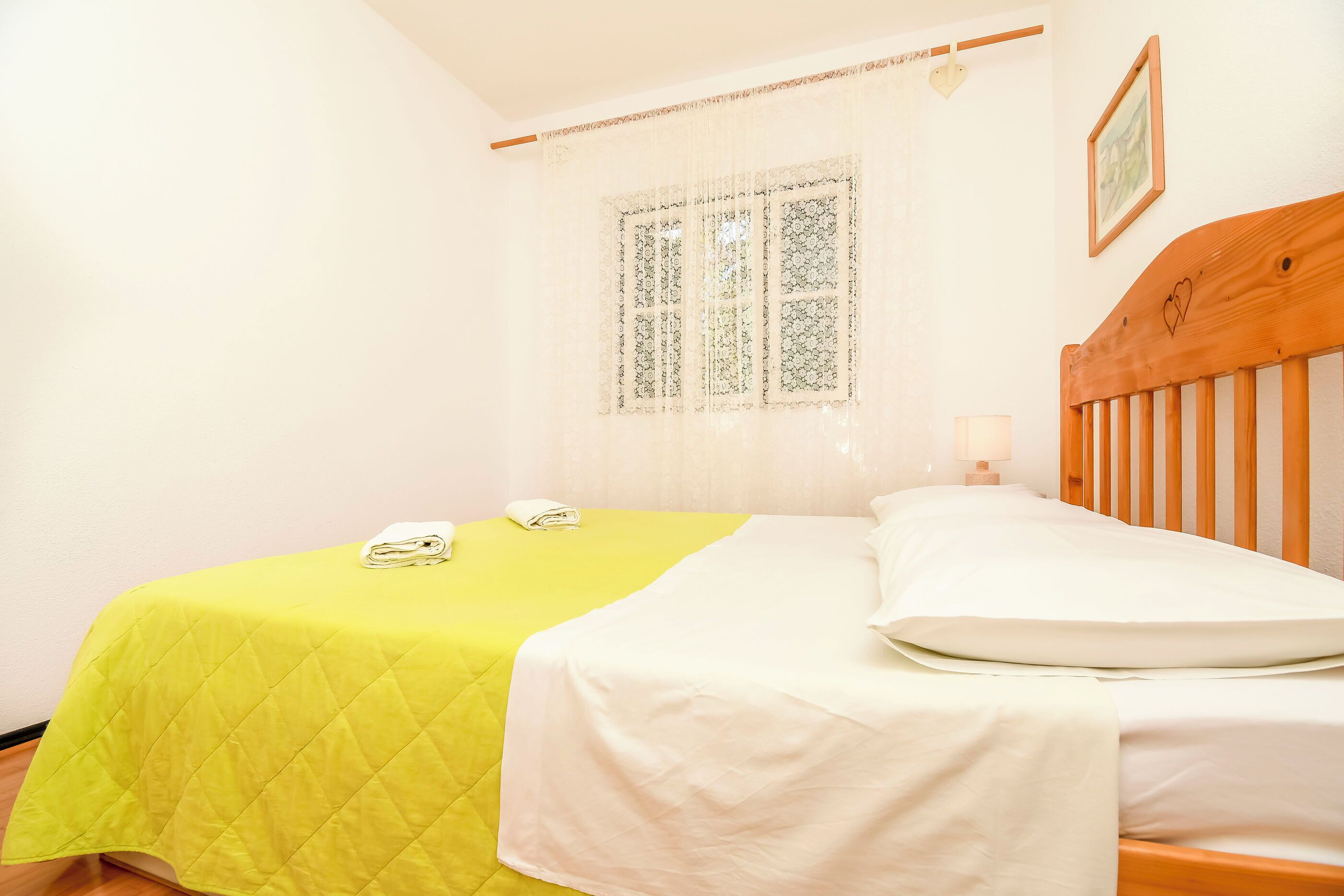 Ein Bett mit gelber Steppdecke und weißer Bettwäsche, ein hölzerner Kopfteil, ein Fenster mit Spitzenvorhängen und eine Nachttischlampe.