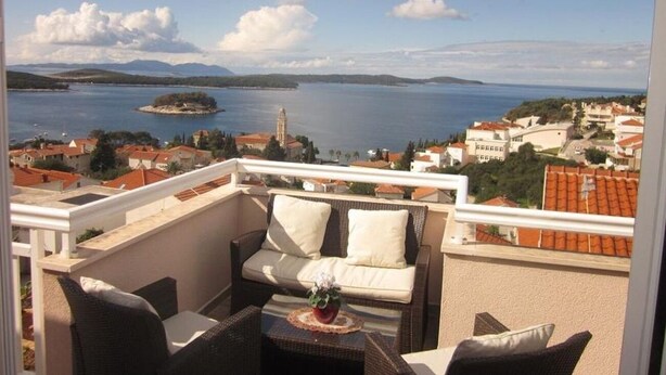 A balcony with a sofa and table, overlooking a lake and town. - Apartments Sto (Hvar)