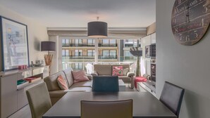 A modern living room with a dining table, sofas, and a large window offering a view of buildings. - 2 bedroom accommodation in Nieuwpoort (Nieuwpoort)
