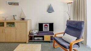 A room with a television displaying 'ERA Real Estate Servivo', a wooden cabinet, a sailboat decoration, a floor lamp, and a modern chair.