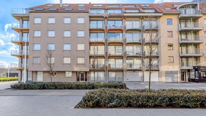 A multi-story residential building with balconies, a central entrance, and a landscaped area with shrubs.