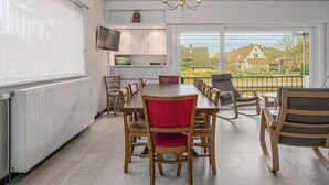 A dining area with a long table, several chairs, a television on the wall, and a view of houses through large windows. - 5 bedroom accommodation in Koksijde (Koksijde)