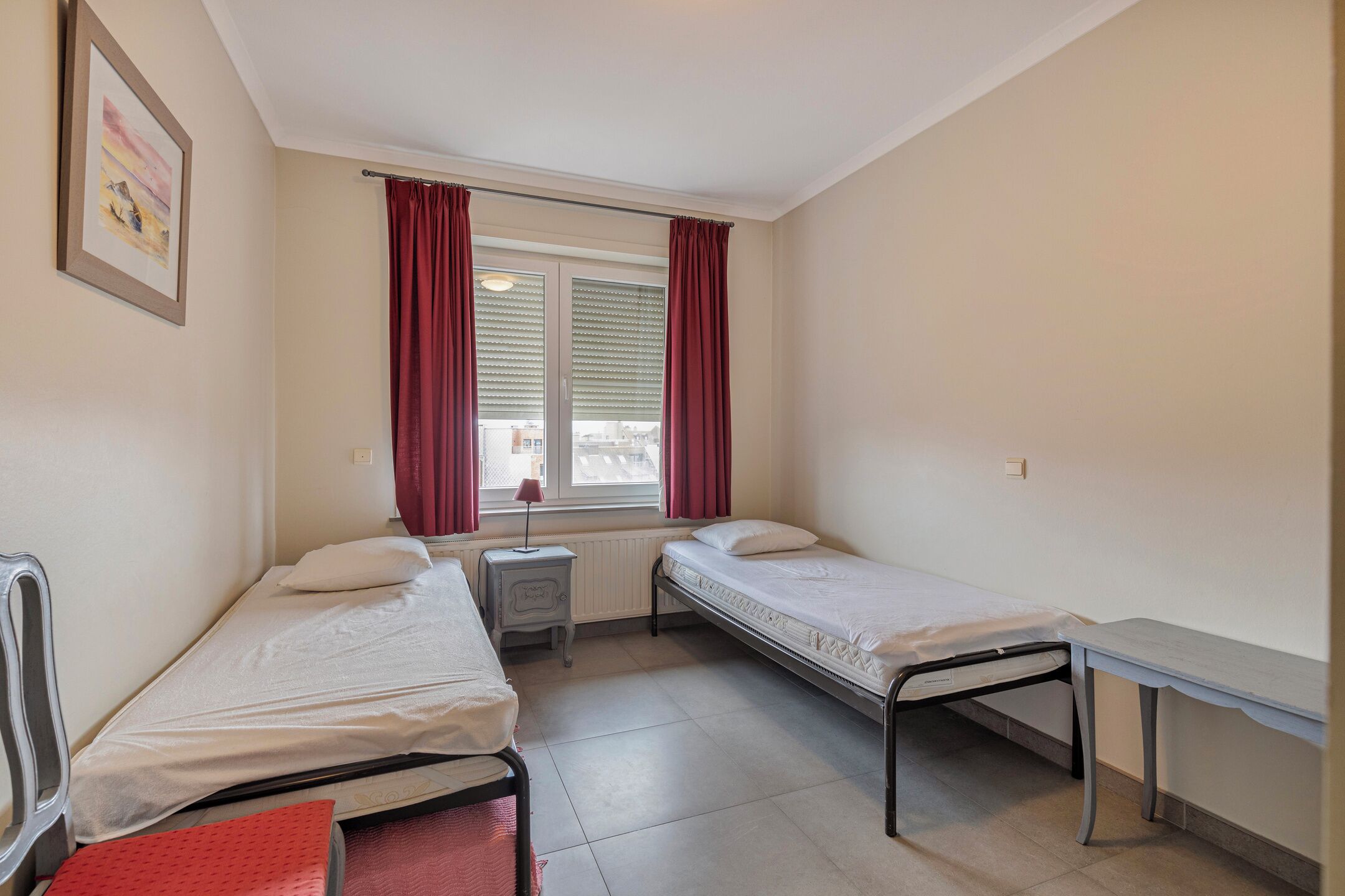 Una habitación pequeña y limpia con dos camas individuales, una mesita de noche, un radiador y una ventana con cortinas.