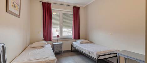 Una habitación pequeña y limpia con dos camas individuales, una mesita de noche, un radiador y una ventana con cortinas.