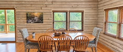 Interior de uma cabana de madeira com uma mesa de jantar rodeada por cadeiras, grandes janelas e um quadro na parede.