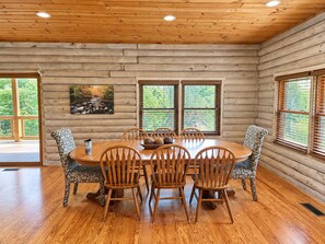 L'interno di una baita di legno con un tavolo da pranzo circondato da sedie, ampie finestre e un quadro appeso al muro.