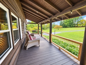 A covered porch with a wooden deck, wicker chairs, and a view of a green landscape.